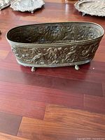 Side view of the oval metal flower pot with ornate embossed decorative patterns and small feet.