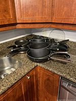 Photo showing several black non-stick frying pans and saucepans stacked on a kitchen granite countertop near a sink with a glass lid visible.