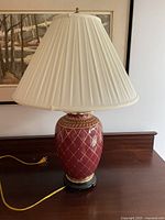Front angled view of the red ceramic lamp with gold lattice pattern and white pleated shade placed on a dark wooden surface.