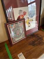 Two framed artwork prints leaning on wall on wooden surface, one smaller indigenous signed print and one larger mandala spiritual print.