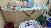 Ironing board with patterned cover, laundry basket, red bucket, and mop in corner