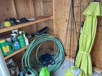 Wide view of garden hoses coiled on floor, fertilizers and sprays on wooden shelves, green garden umbrella and black metal garden trellis in corner