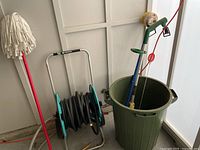 Photo showing mop with red handle, hose reel with green and grey frame holding black garden hose coiled around it, and orange extension cord lying on the floor.