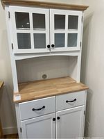 Full view of the white melamine kitchen hutch showing wood-finish top, two glass-paneled doors on upper cabinet, two drawers and lower cabinet doors with black handles and knobs.