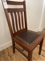 Wooden chair with dark brown riveted leather seat, showing detail of the slatted backrest and seat area with rivets, minor scratches visible on wood frame and leather.