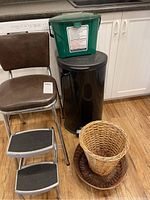 Full lot view showing vintage chair with brown cushioned seat, metal step footrest, black trash bin with green lid, one small wicker basket, and round wicker tray.