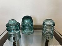 Three antique glass insulators displayed side-by-side on clear surface with white background, showing distinct shapes and embossed detail.