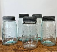 Five canning jars arranged in a line, showing the variation in size and color with blue and clear glass. All jars have metal lids and embossed crown logos.