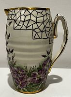 Front-right angled photo of antique ceramic pitcher showing handpainted purple flowers, geometric pattern and gold rim and handle accents.