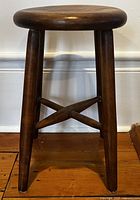 Front view of dark stained wooden milk stool showing round seat and four legs with cross stretchers.