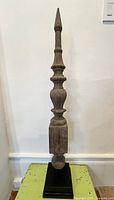 Front view of tall wooden finial standing on black base, showing entire shape and base detail