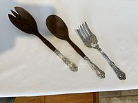 Three serving utensils on white surface showing wooden spoon, wooden fork both with sterling silver handles, and one silver plated fork with detailed handle.