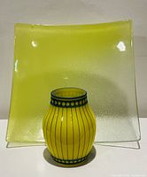 Square yellow fused glass plate with textured surface shown with small yellow and turquoise printed vase in front.