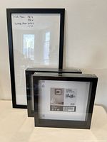 Photo showing two black square frames in original packaging stacked in front of the larger unwrapped frame.