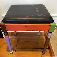 Front view of the handpainted piano stool with black cushioned seat and cat paw prints, showing multiple color blocks on legs and frame.