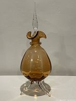 Full view of the amber glass decanter with tall ruffled stopper and three clear glass feet against a plain wall.