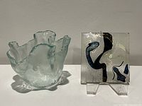 Two fused glass art pieces: a clear wavy-edged vessel and a square glass coaster/trivet with abstract colored patterns, placed side by side on a gray surface against a white backdrop.