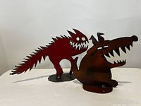Photo of both red cat and burnt orange dog steel sculptures side by side on white surface against white background.