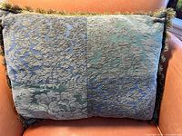 Front side of cushion showing green and blue brocade velvet floral damask pattern with fringe trim.