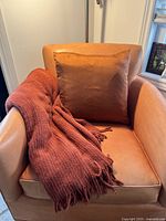 Wide view of the pillow seated on a leather chair with the chenille throw draped alongside. Shows the texture and color of the silk pillow cover and the plush throw.