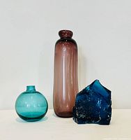 Three vintage glass pieces: two handblown colored vases and one blue slag glass sculpture shown against a plain white background.