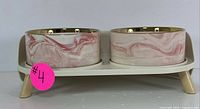 Side view of the set showing two ceramic bowls with pink marble-like design and gold rim placed on beige elevated stand with angled legs.