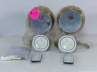 Two wireless LED puck lights wrapped in plastic with remotes and instruction manual laid in front. Bright white LED bulbs visible on puck lights.