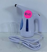 Side view of the handheld steamer with power cord coiled, pink lot number sticker visible on the side. Clear measuring cup placed next to it.