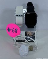 Black smartwatch with attached black band displayed on a clear stand along with a white interchangeable band and a wireless charging dock with USB cable.