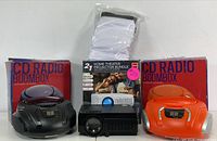 Two CD radio boomboxes (black and orange), a home theater projector, and its foldable white screen displayed on a white surface.