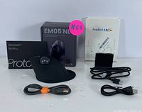 Clear photo of the black ergonomic wireless trackball mouse with USB cable in front, original packaging of mouse and USB adapter box in background, cables laid out neatly.
