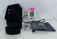 Photo showing digital blood pressure monitor with screen, grey adjustable cuff laid out, pulse oximeter, and three black adjustable support braces grouped on white background.