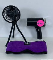 Set of three items: black portable mini fan with flexible legs, compact black hair dryer, and purple heated eye mask laid out on white surface