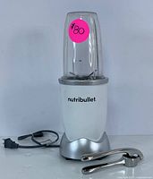 NutriBullet blender with power base, clear blending cup with blade assembly attached, and power cord visible. Small pink sticker number on cup.