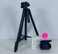 Black adjustable camera tripod standing with Rolosar wireless earbuds charging case and box beside it.