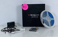 Photo of kit components including Roquati LED strip light reel, black power adapter, white controller unit, and remote control on white background with black Roquati box behind.