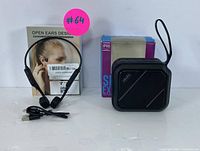 Wireless bone conduction headphones next to the portable Bluetooth speaker with its original box; speaker is black, square-shaped, with control buttons visible and a black rubber strap for carrying.