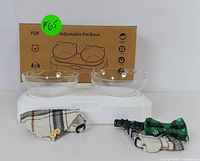 Photo of adjustable pet bowl with two clear bowls on white base, two plaid bandanas, and green bow tie collar with black and white pattern in front. Box in background with product name.