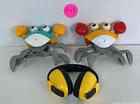 Two interactive crawling crab toys, one with blue and yellow claws and the other with yellow and red claws, along with a pair of yellow and black noise-reduction headphones.