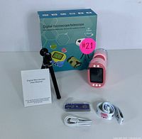 Digital microscope with tripod, user manual, USB charging cable, blue plastic accessory, and lanyard arranged around the original box.