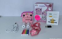 Kids pink instant print camera with face design, two rolls of instant print paper, USB charging cable, six color markers, pink unicorn-themed lanyard, USB receiver, and battery precautions leaflet laid out on white background.