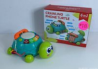 Front angle of crawling phone turtle toy alongside box showing 6-in-1 features such as phone handset, musical and learning buttons.
