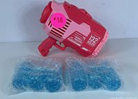 Red and pink electric gel ball blaster with two packs of blue gel balls wrapped in bubble wrap placed in front on white background