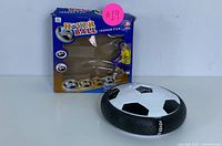 Front side view of the Hover Ball Indoor Soccer Toy in black and white soccer ball design including its original blue and orange box.