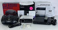 Photo showing Sylvania dual screen portable DVD player box and unit, Proscan HDMI upconvert DVD player with remote and power cord, compact CD radio boombox, and white multimedia projector with remote and accessories.
