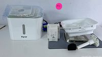 Photo of all items including Parner automatic water fountain, digital pet scale, filter parts laid out on white table against white background.