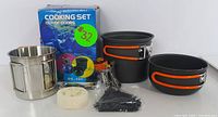 DS-101 cooking set showing two black non-stick pots with orange foldable handles, stainless steel cup, small compact stove burner, white cleaning sponge and accessories in front of original box with green price sticker.