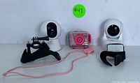 Two white smart security cameras with wrist straps alongside a pink kids waterproof camera with clear protective casing, placed on a white surface.