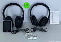 Front view of two black wireless over-ear headphones alongside a compact MP3 player with a color display screen and three charging cables; includes the MP3 player user manual.