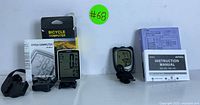 Photo showing bicycle computer with display, mounting parts, instruction sheet, and packaging alongside digital stopwatch and instruction manuals